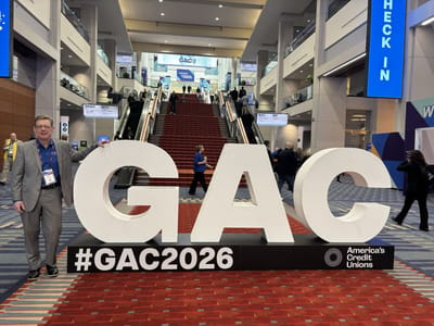 Greg Cook standing beside giant white GAC letters with a #GAC2026 America's Credit Unions sign in the convention center lobby, escalators and attendees behind him
