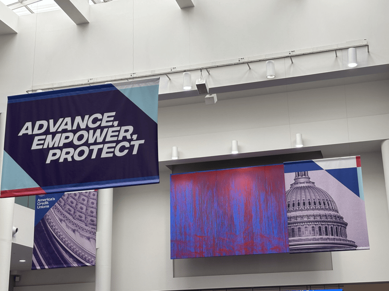 Convention center banners reading ADVANCE, EMPOWER, PROTECT alongside America's Credit Unions banners showing the U.S. Capitol dome
